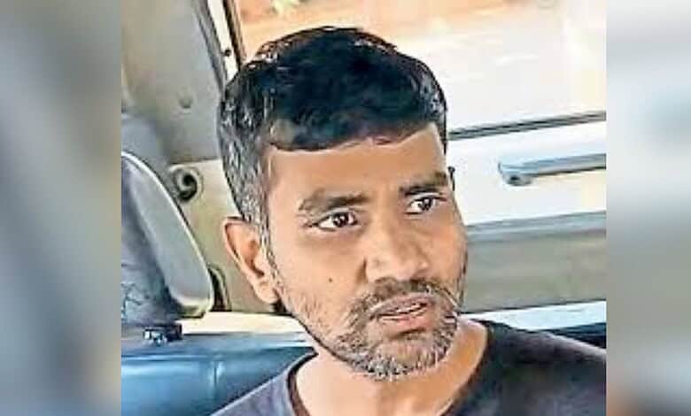 Man with short black hair and beard sitting in a vehicle, looking serious.