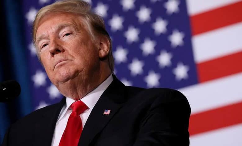 Donald Trump speaking at a press event with US flags behind him.