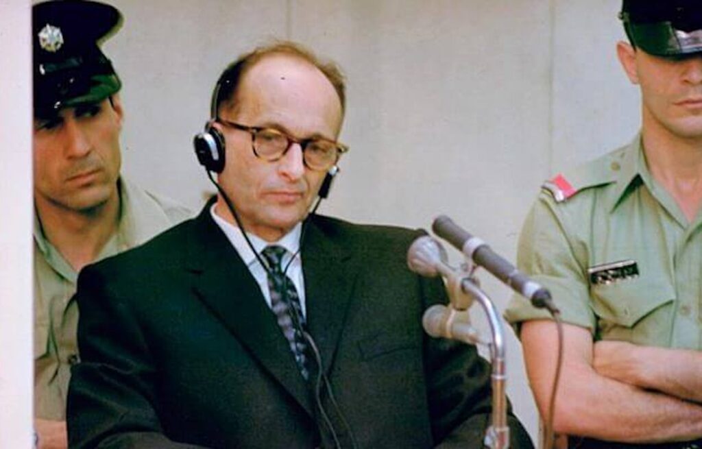 Adolf Eichmann listens through headphones during his 1961 trial in Jerusalem, guarded by Israeli officers.