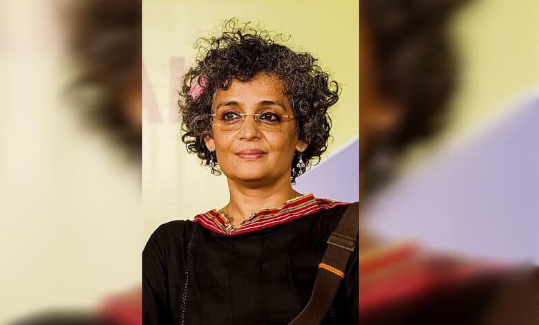 Arundhati Roy at a public event, wearing glasses and a black outfit with a red-bordered neckline.