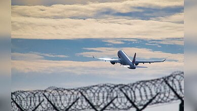 Image shows a plane in the sky above a barbed wire fence, representing themes of deportation and seeking safety.