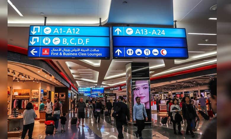 Passengers walk through a departure terminal at Dubai International Airport with gate signs overhead.