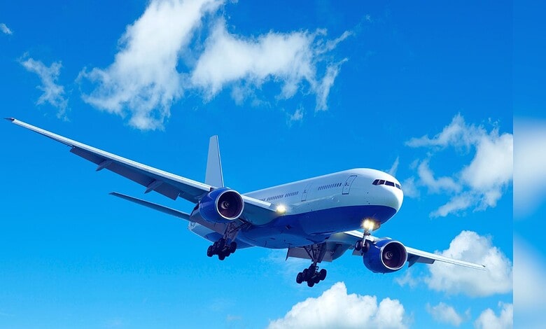 Image shows A large commercial airplane with landing gear deployed flies through a bright blue sky with scattered white clouds.