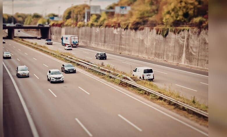 Image shows Multi-lane highway with light traffic including cars, trucks, and vans traveling in both directions, bordered by concrete retaining walls and vegetation with a tilt-shift photography effect.