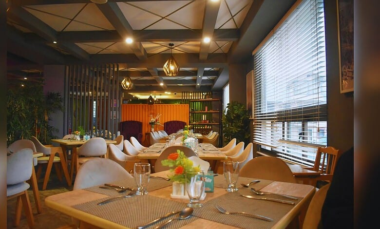 Modern restaurant interior featuring warm wood tables with white chairs, geometric coffered ceiling, and natural light from large windows