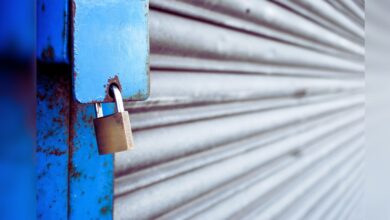 Image shows A brass padlock secures a weathered blue metal shutter with visible rust and peeling paint.