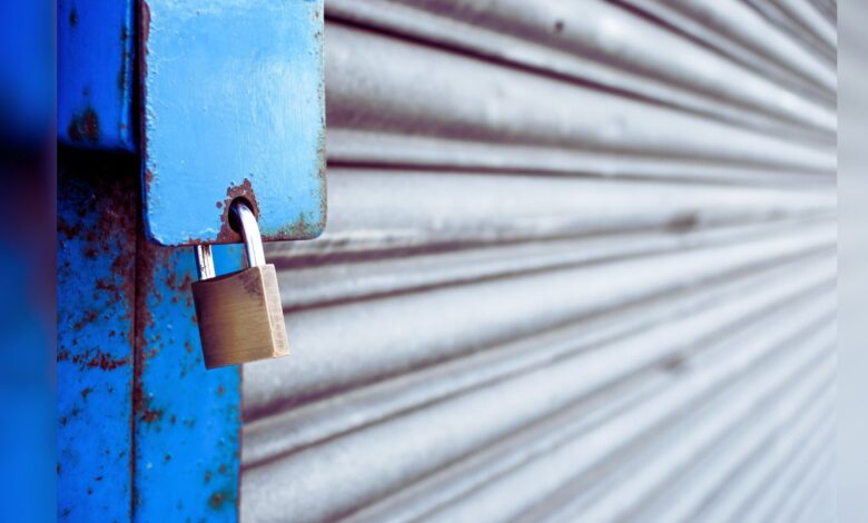 Image shows A brass padlock secures a weathered blue metal shutter with visible rust and peeling paint.