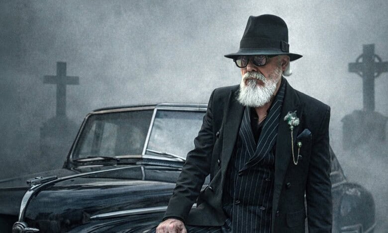 Elderly man with white beard, sunglasses, and black hat standing beside vintage car in foggy graveyard.
