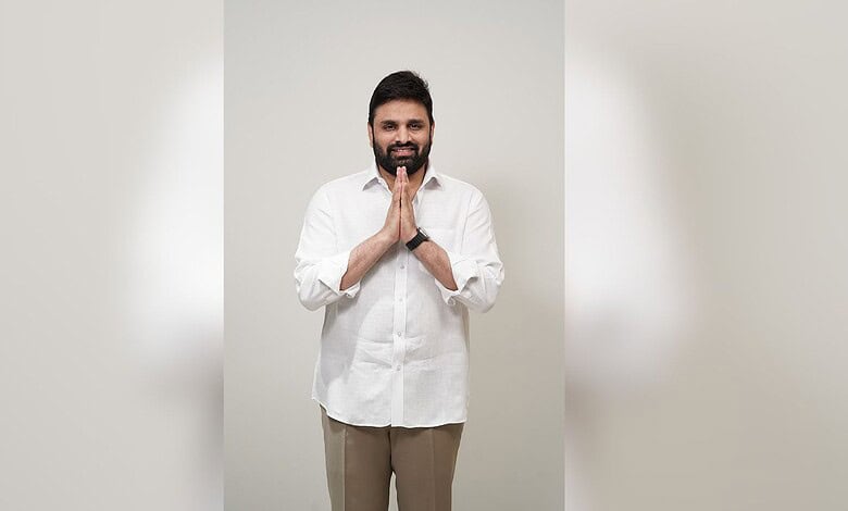 An image of a smiling man with hands folded in a gesture of greeting or respect, standing against a plain.