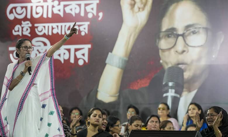 Mamata Banerjee addressing a rally, gesturing with her hand, with a large poster of herself behind her.