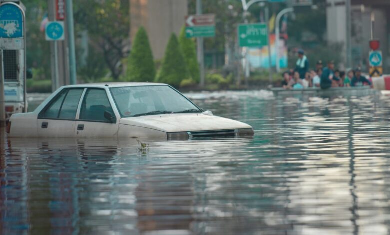 Image shows A white car nearly submerged in floodwater on a city street as onlookers watch from higher ground nearby.