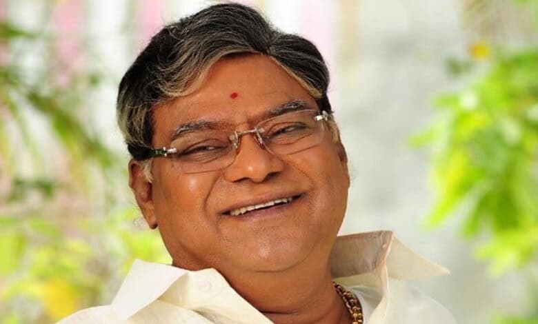 An elderly Indian man smiling warmly, wearing traditional attire and glasses, in a bright, natural settin.
