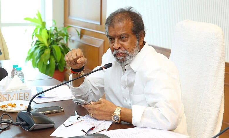 An elderly man speaking at a meeting about Telangana's public healthcare system reform, seated at a desk.