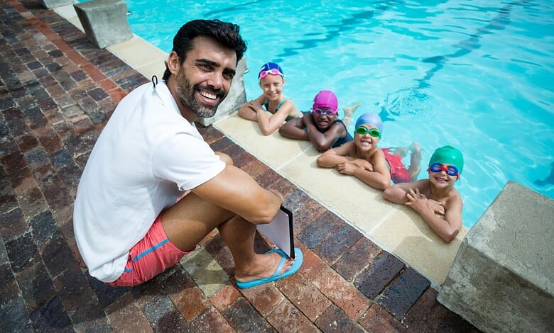 Male swimming coach with four children in goggles at the poolside during summer sports coaching camp.