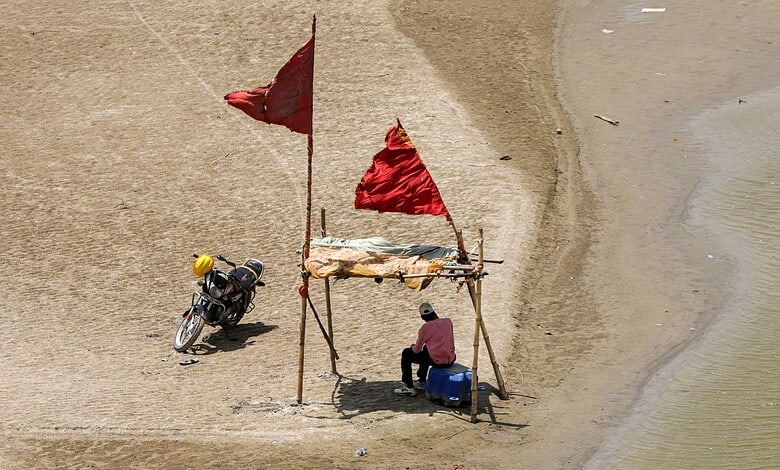 Small makeshift shelter on a sandy beach with a person sitting under it, near water, during a heatwave.