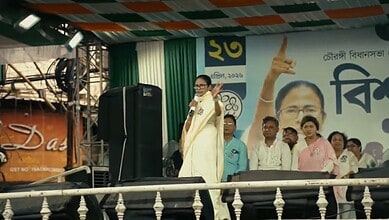Politician speaking at a public rally with supporters and a large poster in the background.