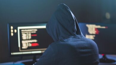 Image shows Person in a hoodie working on a computer with code on multiple screens in a dark setting