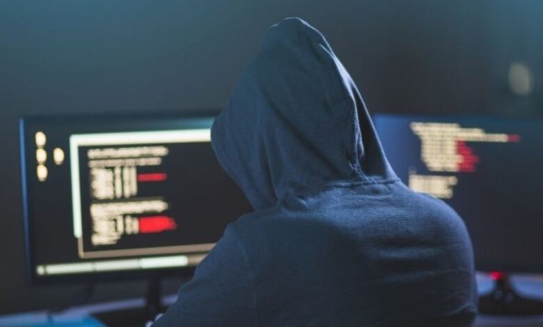 Image shows Person in a hoodie working on a computer with code on multiple screens in a dark setting