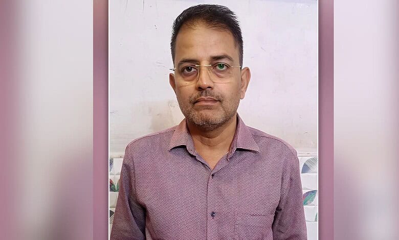 Man with glasses wearing a casual shirt, standing indoors against a plain background.