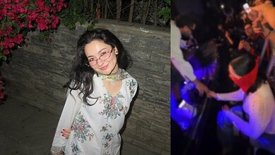 Hania Aamir smiling at a social gathering, wearing glasses and a floral dress, with a lively background.