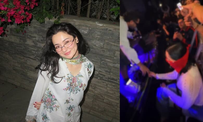 Hania Aamir smiling at a social gathering, wearing glasses and a floral dress, with a lively background.