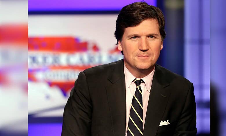 Tucker Carlson poses in a television studio wearing a dark suit and striped tie.