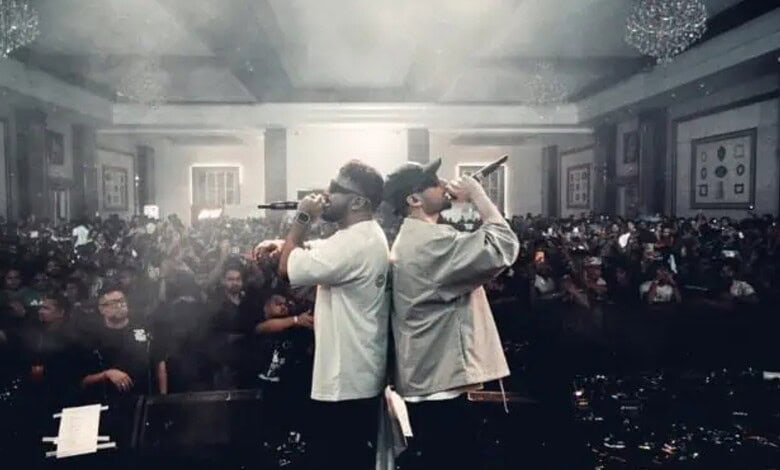 Two hip-hop artists performing on stage at 'LEGACY' show with a large audience in the background.