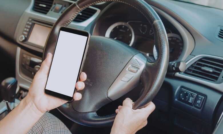 Image shows A driver holding a smartphone with a blank screen while steering a car with their other hand.