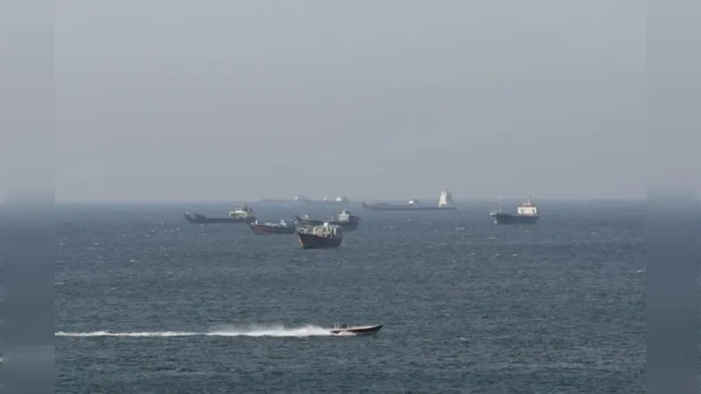 Cargo ships anchored in the Strait of Hormuz with a speedboat passing during shipping disruption.