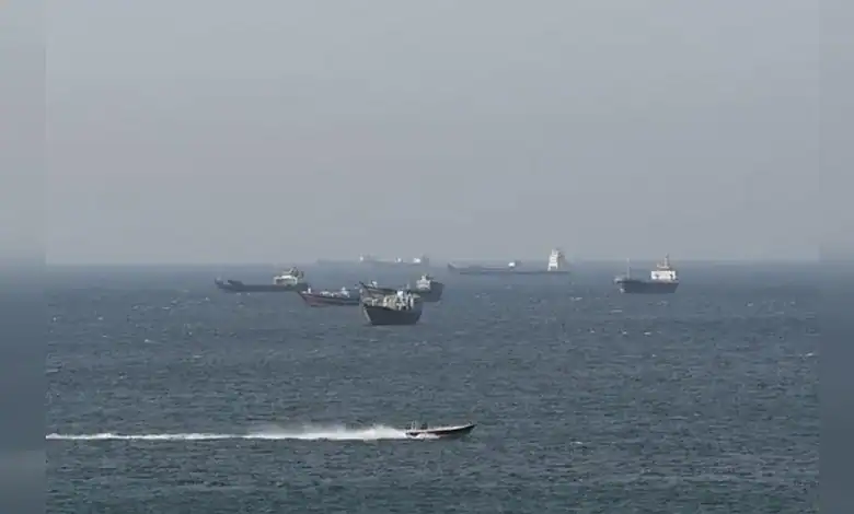Cargo ships anchored in the Strait of Hormuz with a speedboat passing during shipping disruption.