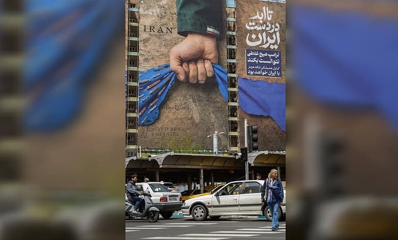 Hand gripping blue fabric in front of a large billboard in Iran, with cars and pedestrians nearby.