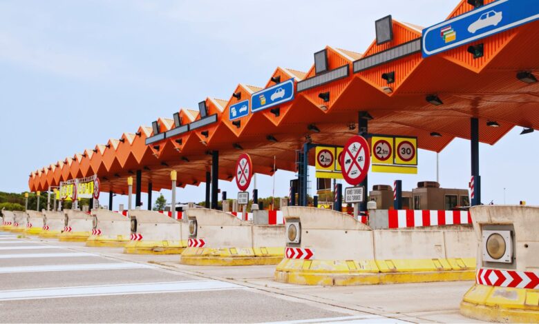 Image shows A multi-lane highway toll plaza with a distinctive orange zigzag canopy roof, traffic signs, and concrete lane dividers.