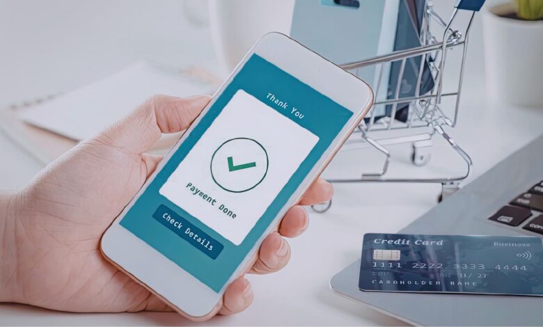 Image shows A hand holds a smartphone showing a "Payment Done" confirmation screen, with a credit card and miniature shopping cart on a desk nearby.