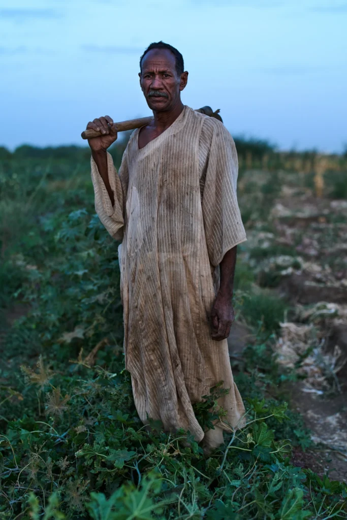 Sudan farmer standing in a field holding a farming tool, highlighting agricultural struggles caused by gl.