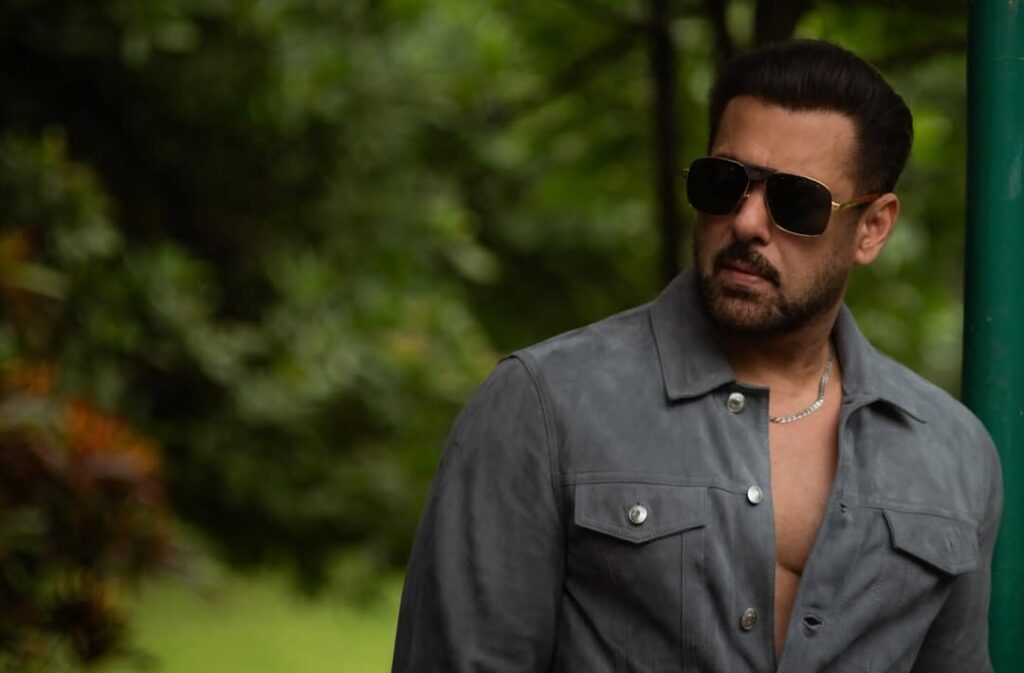 Man wearing sunglasses and a grey jacket in a lush green park setting.