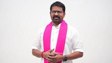 A man wearing glasses and a white shirt with a pink scarf, standing against a plain background.