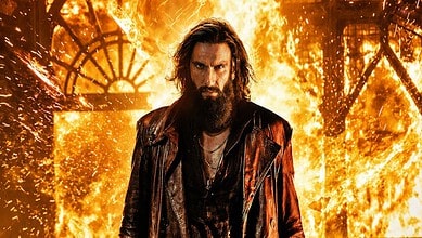 A man with long hair and a beard standing in front of a fiery background with sparks and flames.