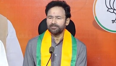 A woman with glasses and a beard, wearing a grey kurta and a yellow-green scarf, speaking at a press conf.