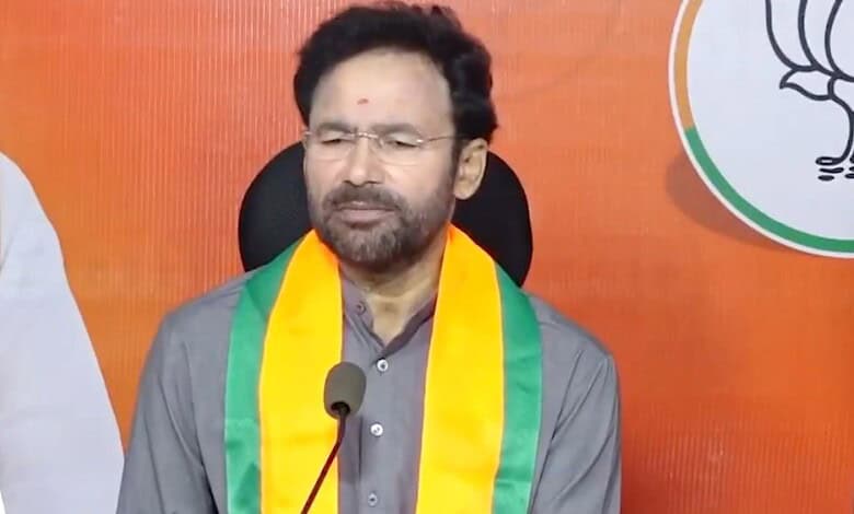 A woman with glasses and a beard, wearing a grey kurta and a yellow-green scarf, speaking at a press conf.