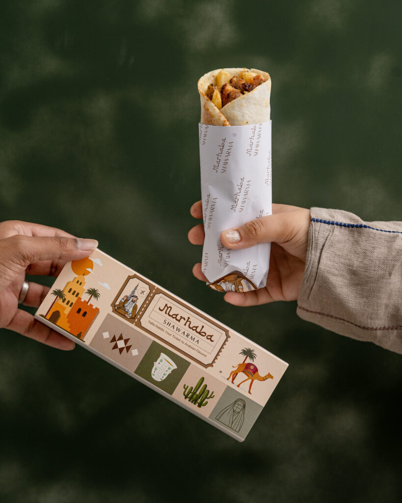 Close-up of a hand holding a wrapped shawarma and a branded Siasat Daily card against a blurred green bac.