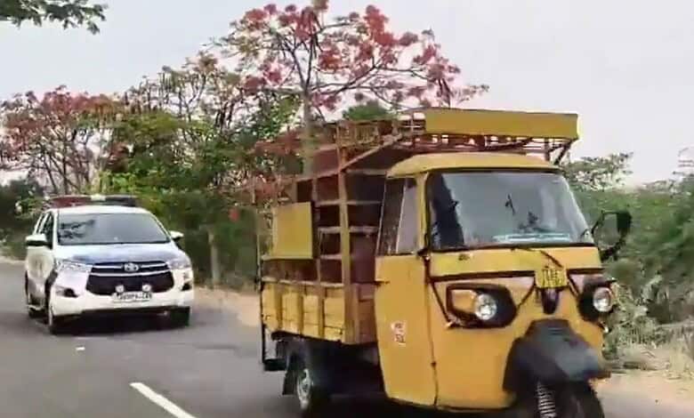 Yellow auto rickshaw on a rural road with a white car in the background, greenery and pink flowering tree.
