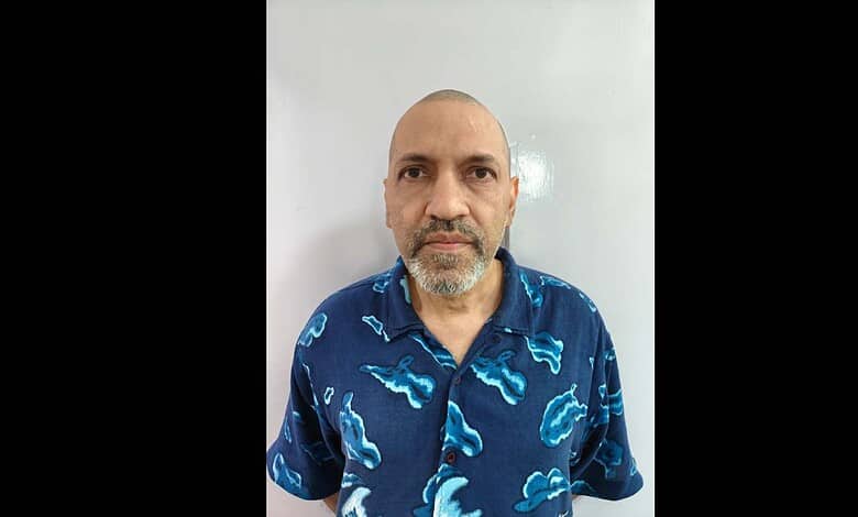 Man with a beard and shaved head, wearing a blue shirt with cloud patterns, standing against a plain back.