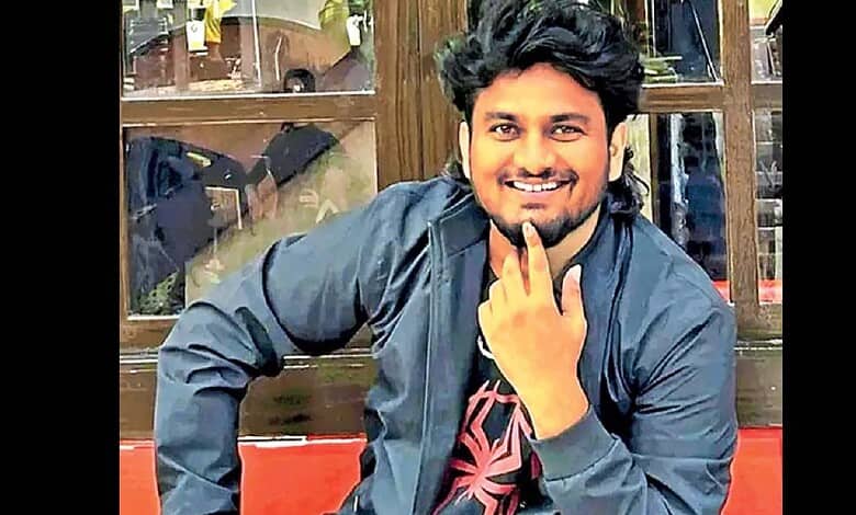 A smiling young man with dark hair and a beard, wearing a black jacket, sitting indoors in front of a woo.