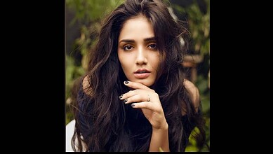 A woman with long, wavy dark hair posing outdoors in a natural setting.