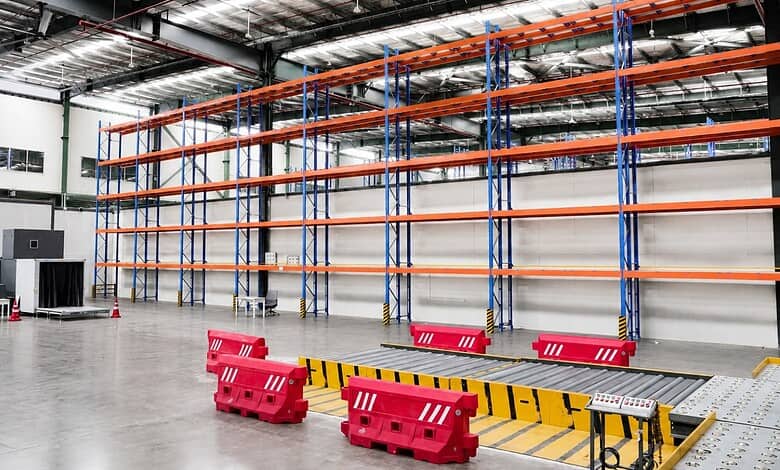 Aisle in a large warehouse with empty blue and orange shelving units and red safety barriers on the floor.