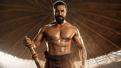 A muscular man with a beard holding a wooden stick in a traditional setting.