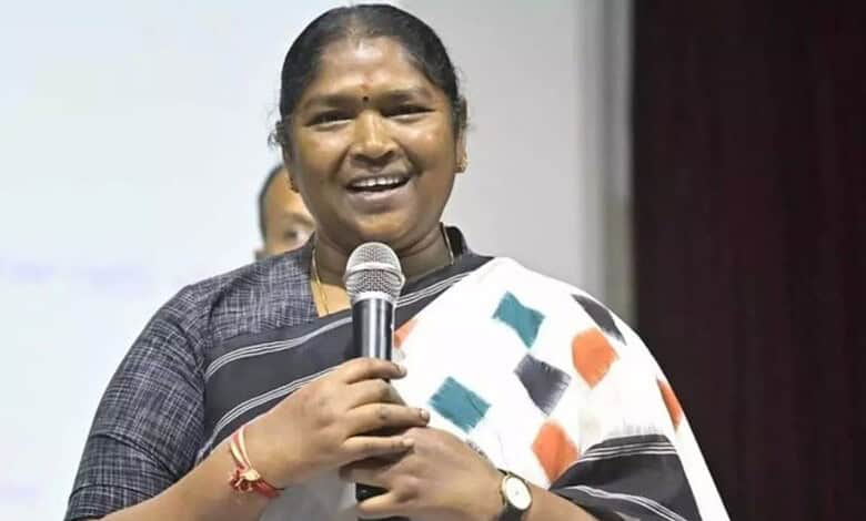Woman speaking into microphone at a press event, wearing a saree with a black and white pattern.