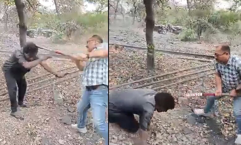 Two men fighting with sticks in a wooded area, one man in a checkered shirt and sunglasses, the other in.
