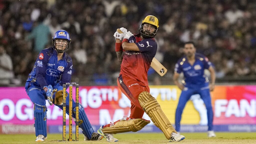 RCB player hits the ball during a tense IPL match against MI, with players and crowd in the background.
