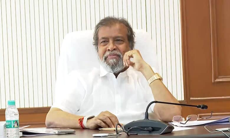 A man with grey hair and beard sitting at a desk in an office setting, wearing a white shirt and a gold w.